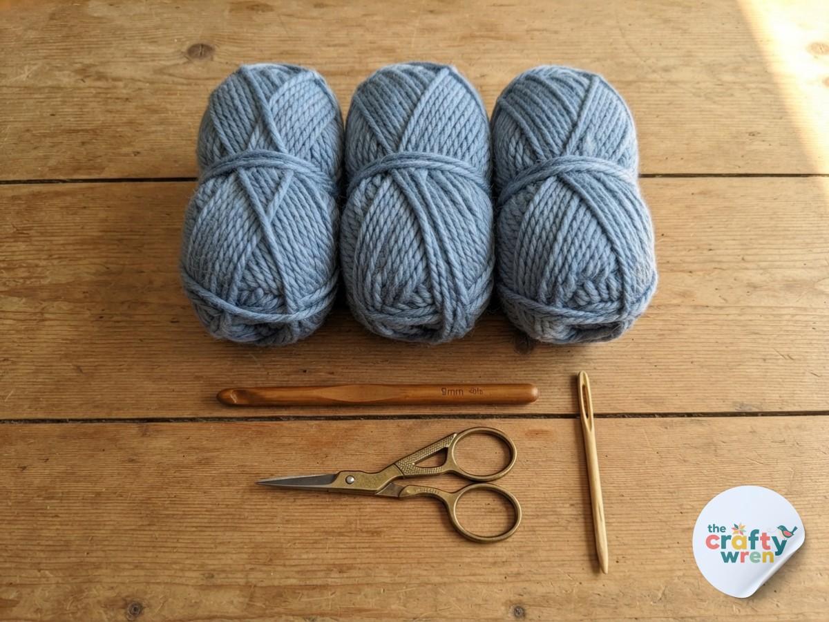 Chunky dusty blue yarn, wooden hook, and scissors ready to start an easy textured crochet blanket project.